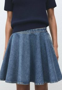 Une personne portant un haut en tricot bleu foncé rentré dans une jupe en denim bleu à taille haute et évasée, se tenant contre un mur blanc.