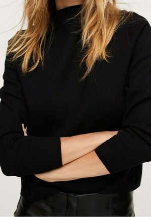 Femme aux longs cheveux blonds portant un haut à manches longues noir et un pantalon noir, les bras croisés au niveau de la taille, sur un fond uni.