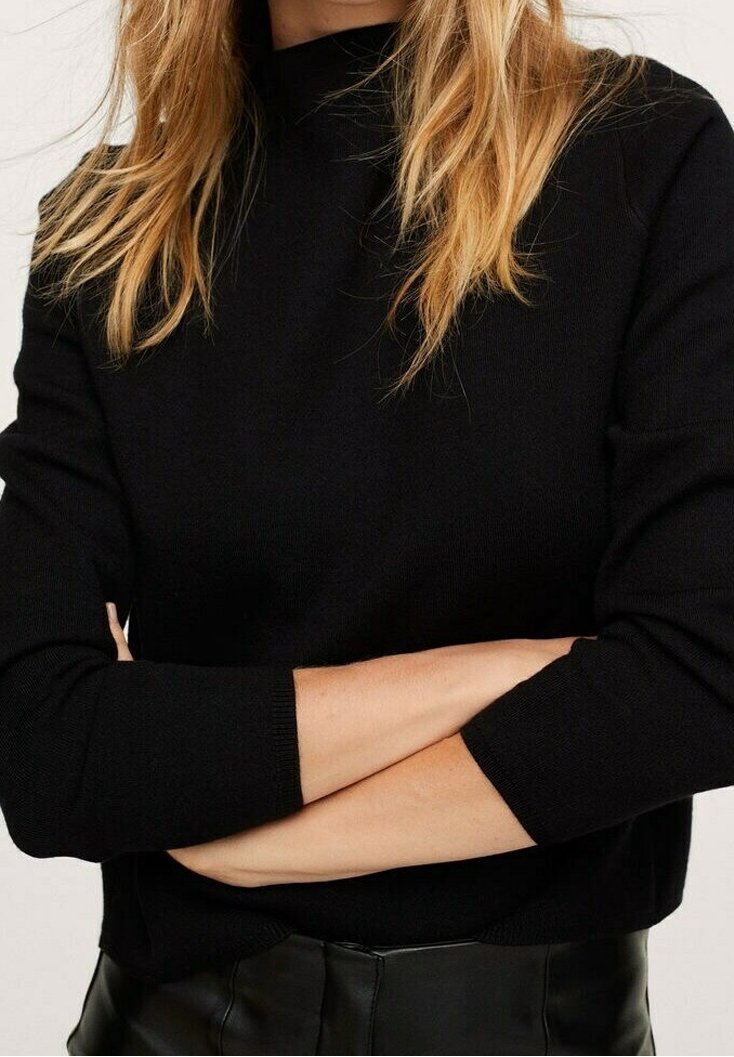 Femme aux longs cheveux blonds portant un haut à manches longues noir et un pantalon noir, les bras croisés au niveau de la taille, sur un fond uni.