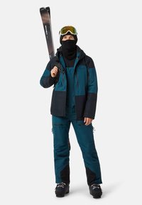 Uomo vestito con tuta da sci blu e nera, con occhiali da sci e passamontagna, che tiene gli sci in verticale sulla spalla, in piedi su sfondo neutro.