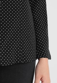 Czarny, długi rękaw bluzka w białe kropki, z gładką teksturą i lekko rozkloszowanymi mankietami. Widoczne detale szwów po bokach.