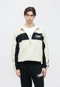Αμάνικο μπουφάν Nike σε εκρού και μαύρο χρώμα, με φερμουάρ μπροστά, ελαστικά μανίκια και κεντημένο λογότυπο. Συνδυάζεται με μαύρο παντελόνι φούτερ.