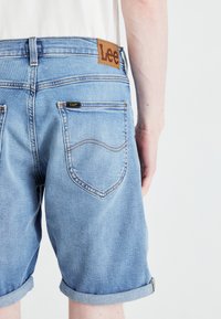 Ανοιχτό μπλε τζιν σορτς με διπλωμένα μπατζάκια, με καφέ δερμάτινη ετικέτα στη πίσω ζώνη και αντιθετικές ραφές στις τσέπες.