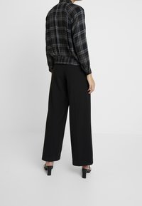Schwarze karierte Bluse mit elastischem Saum, langen Ärmeln und taillierten Manschetten; kombiniert mit weit geschnittenen schwarzen Hosen und schwarzen Absatzsandalen.