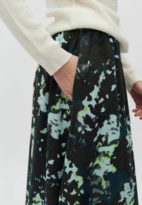 Osoba nosící krémový žebrovaný svetr s rukou v kapse plisované sukně s abstraktním květinovým vzorem v černé, zelené a modré barvě.