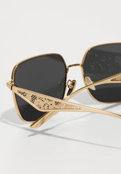 Gafas de sol doradas con lentes cuadrados negros, que presentan intrincados patrones recortados en las patillas y una marca grabada en el brazo.