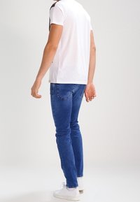 Pánské bílé tričko s černými akcenty, spárované s modrými úzkými džíny a bílými teniskami, ukazující čistý, moderní design a střih.