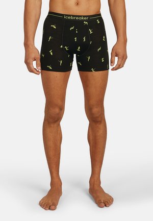 Uomo in boxer neri con silhouette di sciatori gialli e logo "icebreaker" nella cintura, in piedi scalzo su uno sfondo semplice.