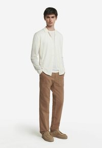 Vit knappad kofta över en vit t-shirt, kombinerad med bruna, rymliga byxor och beige mockaskor. Tydlig design och neutrala färger.