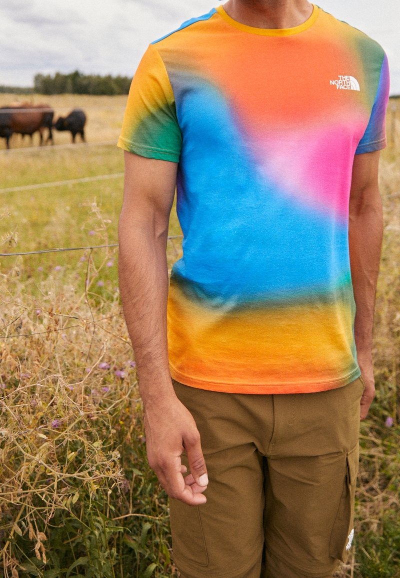 Muž v barevném tričku s tie-dye od značky The North Face a hnědých cargo šortkách stojí na travnatém poli s krávami v pozadí.