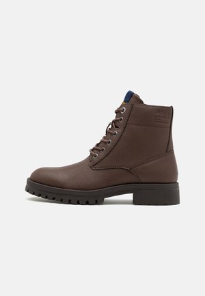 Bota de ante marrón con cordones, suela resistente y costuras tonales, que presenta un cuello acolchado y un logotipo de la marca en relieve en el lateral.
