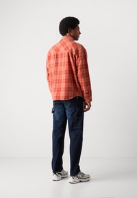 Camisa a cuadros roja y naranja con mangas largas, puños abotonados, combinada con pantalones de mezclilla azul oscuro y zapatillas grises. Estilizada desde la parte posterior.