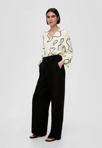 Witte blouse met zwart abstract lijnpatroon, V-hals, lange mouwen, gecombineerd met hoge zwarte broeken en zwarte sandaaltjes met bandjes.