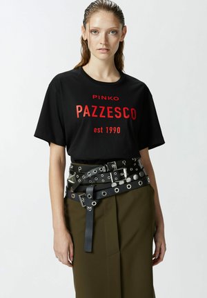 Crna pamučna majica s crvenim otiskom "PINKO PAZZESCO est 1990." Uparena s višenamenskim crnim remenom koji ima okrugle rupice i srebrne detalje.