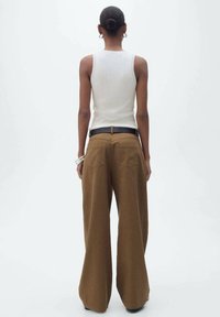 Braune, weit geschnittene Hose mit zwei Gesäßtaschen, getragen mit einem engen weißen Tanktop und einem schwarzen Gürtel. Glatte Stofftextur, minimalistisches Design.