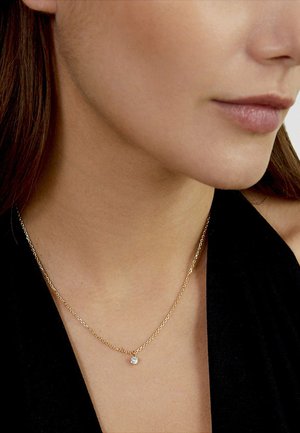 Nahaufnahme des Halses einer Frau, die eine filigrane goldene Halskette mit einem einzigen kleinen runden Diamanten-Anhänger trägt.