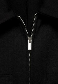Sort zip-up jakke med en sølvmetal lynlås, tekstureret stof og to applikerede lommer foran. Glat krave og syningsdetaljer.