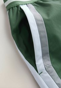 Tissu vert avec des rayures verticales blanches et grises, montrant un gros plan d'une ceinture froncée et un détail de couture latérale sur un vêtement.