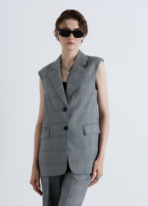 Person wearing a gray checkered sleeveless blazer and matching pants with black sunglasses and a silver necklace against a plain background.