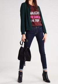 Grøn bomberjakke over en rød sweater med hvid og lyserød tekst, båret med mørkeblå jeans og sorte ankelstøvler, mens der holdes en sort taske.