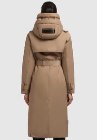 Lunga giacca imbottita beige con colletto alto, vita arricciata e fessure laterali. Presenta una patch con logo sul retro e polsini delle maniche regolabili.