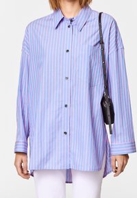Light blue shirt with vertical pink stripes, long sleeves, button closure, and a chest pocket. Paired with a black crossbody bag.