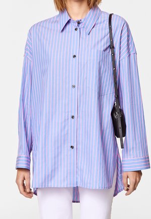 Camicia azzurra chiara con strisce rosa verticali, maniche lunghe, chiusura a bottoni e taschino sul petto. Abbinata a una borsa a tracolla nera.