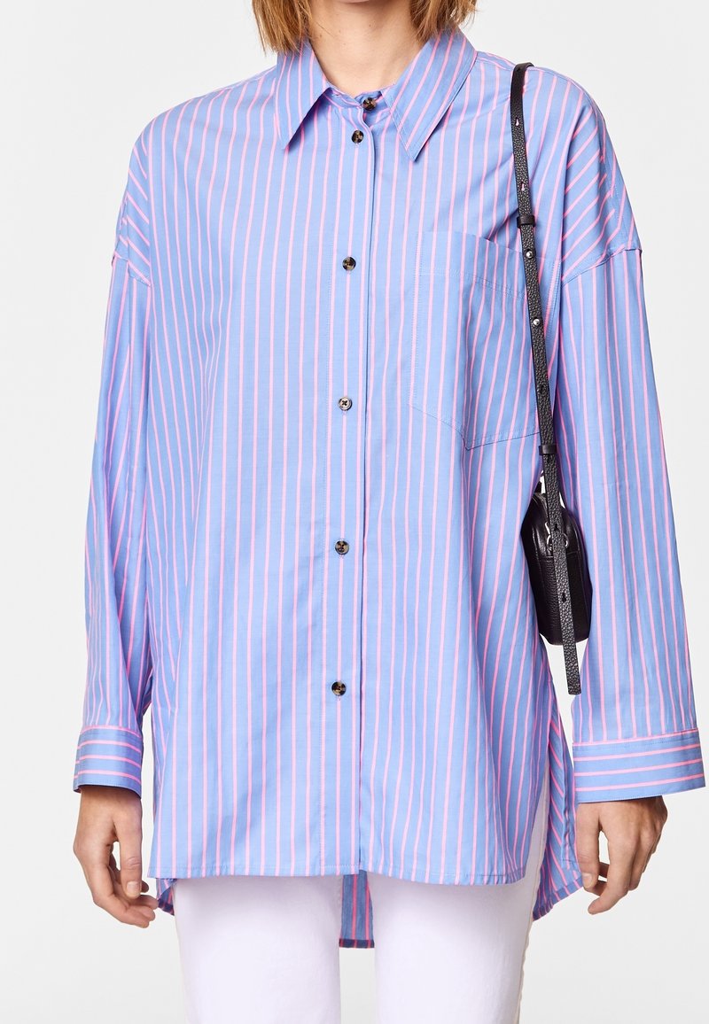 Light blue shirt with vertical pink stripes, long sleeves, button closure, and a chest pocket. Paired with a black crossbody bag.