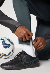 Ciemnoszare spodnie sportowe o teksturowanym wykończeniu, z regulującym zamkiem przy kostce, w połączeniu z czarnymi butami Nike i białymi skarpetami, obok kolorowa piłka nożna.