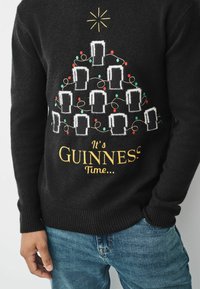 Čierny sveter s vianočným dizajnom stromčeka z pivných pohárov, zdobený farebnými svetlami, s nápisom "Je čas na GUINNESS..." vytlačeným nižšie.