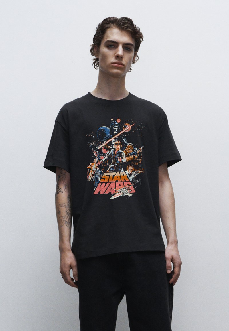 Černé bavlněné tričko s barevným potiskem "Hvězdné války" zobrazujícím postavy a lodě, s uvolněným střihem a krátkými rukávy.