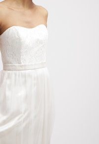 Abito bianco senza spalline con un corpetto in pizzo, una cintura in satin liscia e una gonna trasparente a strati. Il tessuto è leggero e fluttuante.