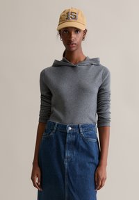 Szara bluza z prążkowaną strukturą i kapturem z kominem, zestawiona z niebieską spódnicą jeansową z wysokim stanem. Modelka ma na sobie żółtą czapkę z numerem 15.