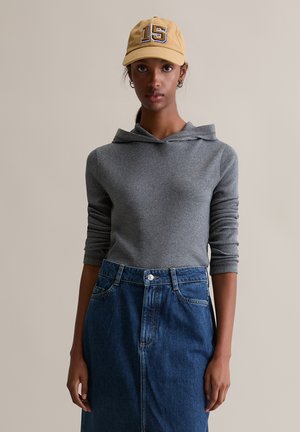 Szara bluza z prążkowaną strukturą i kapturem z kominem, zestawiona z niebieską spódnicą jeansową z wysokim stanem. Modelka ma na sobie żółtą czapkę z numerem 15.