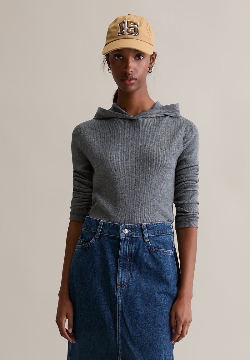 Szara bluza z prążkowaną strukturą i kapturem z kominem, zestawiona z niebieską spódnicą jeansową z wysokim stanem. Modelka ma na sobie żółtą czapkę z numerem 15.