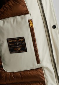 Jakna s punjenjem koja ima kremastu vanjštinu s smešnim akcentima, kviltanu teksturu i žuti zatvarač. Oznaka kaže "NIGHTFLIGHT ISSUE 508/13."