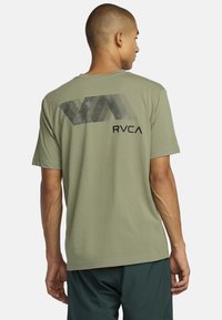 Olive zielona bawełniana koszulka z dużym czarnym logo w formie linii z napisem „RVCA”, krótki rękaw i luźny krój. Widok z tyłu.