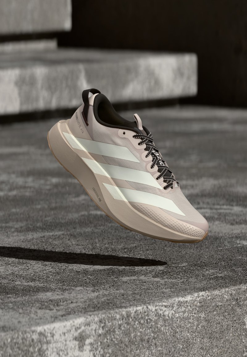 Chaussure de course beige clair avec trois bandes blanches et des lacets noirs lévitant au-dessus d'une surface en béton grise et des marches.