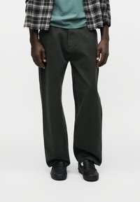 Άτομο που φοράει φαρδιά μαύρη παντελόνια, μαύρα παπούτσια, τιρκουάζ πουκάμισο και ασπρόμαυρο καρό μπουφάν, στέκεται μπροστά σε απλό φόντο.