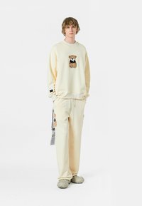 Conjunto de sudadera y pantalones color crema, con un bordado de oso en la parte delantera y lateral. Textura suave y corte relajado. Calzado estilo croc.