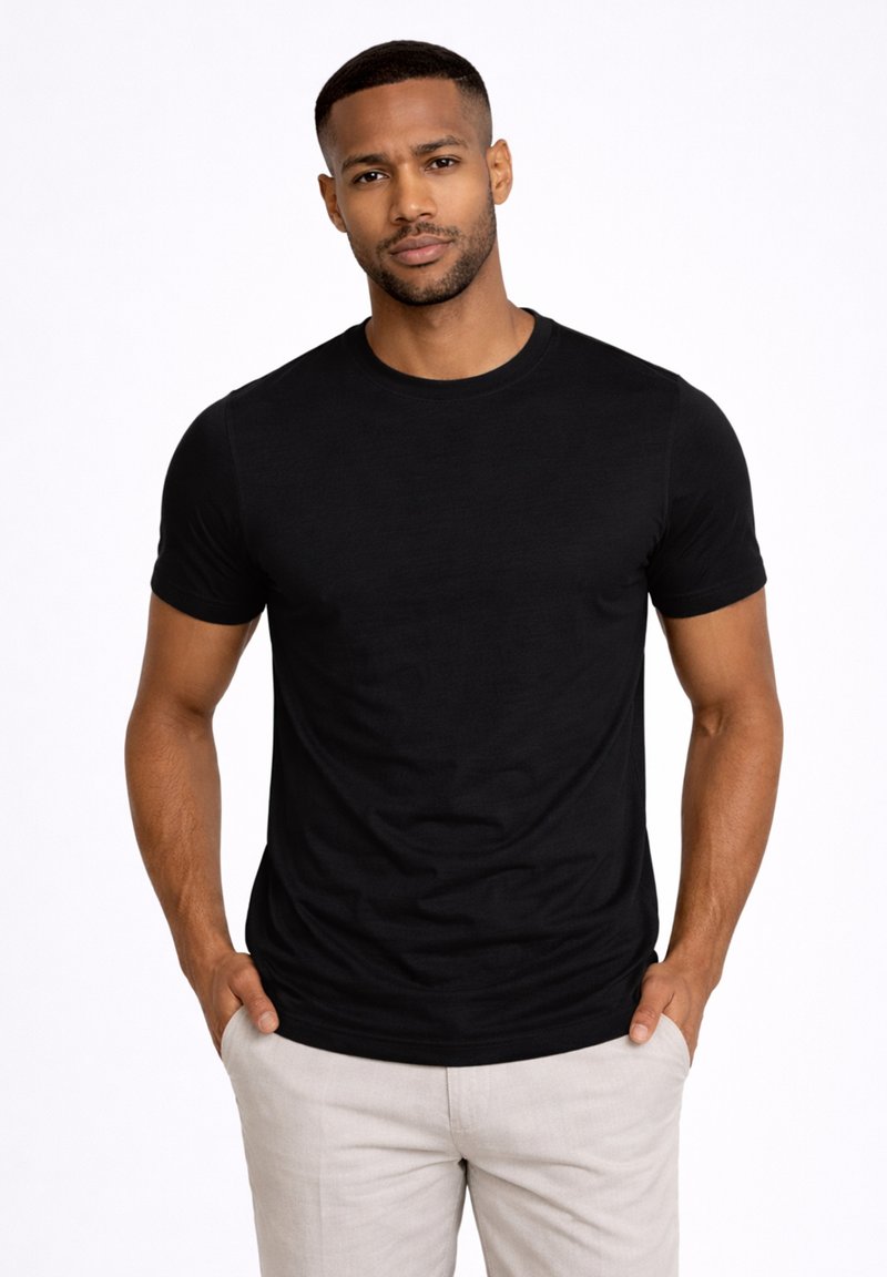 Man met kort haar en baard, gekleed in een zwart T-shirt met korte mouwen en lichtbeige broek, met handen in de zakken.