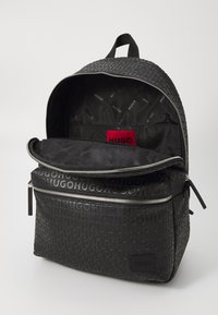 Mochila negra con una superficie texturizada que presenta el logo repetido "HUGO". Tiene un gran compartimento principal, un bolsillo frontal con cremallera y un asa en la parte superior.