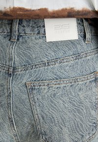 Área del bolsillo trasero de unos pantalones vaqueros azul desgastado con costuras visibles y una etiqueta blanca con la leyenda "GESTUZ" en la cinturilla.