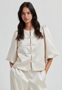 Krémově zbarvená halenka s kulatým výstřihem, nafouklými rukávy a ozdobnými zapínáními vpředu. Hladký, saténový materiál.