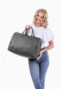 Šedá kožená sportovní taška s hladkou texturou, pevnými držadly a jemným logem na přední straně. Má zaoblené hrany a prostorný design.