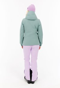 Zelená bunda s kapucí, elastické manžety; v kombinaci se světle fialovými kalhotami. Oblečení má sportovní design vhodný pro zimní aktivity.