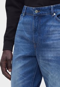 Gros plan d'une personne portant un jean en denim bleu moyen avec un bouton argenté et une chemise noire à manches longues, main reposant à côté de la cuisse.