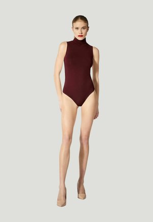 Frau trägt einen ärmellosen hochgeschlossenen burgunderfarbenen Body und beige High Heels, steht vor einem schlichten hellgrauen Hintergrund.