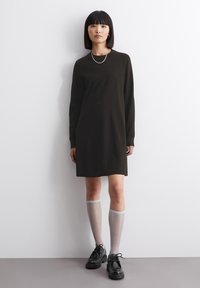 Vestito lungo a maniche lunghe verde scuro con taglio dritto, abbinato a calze grigio chiaro fino al ginocchio e scarpe nere con suola spessa. Tessuto liscio, design minimalista.