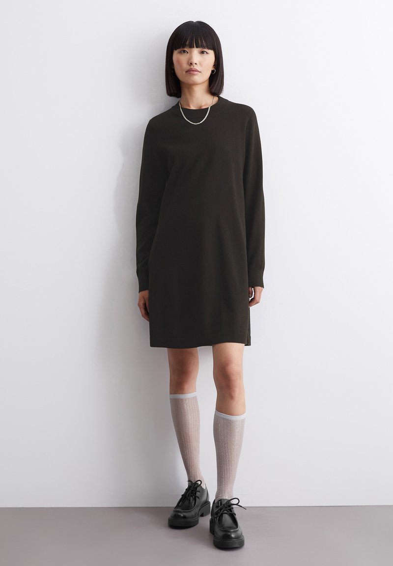 Dunkelgrünes langärmeliges Kleid mit geradem Schnitt, kombiniert mit knielangen hellgrauen Socken und schwarzen klobigen Schuhen. Glatte Textur, minimalistisches Design.